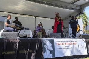 a woman in a long red coat and jazz band performing on stage over a Billie Holiday banner