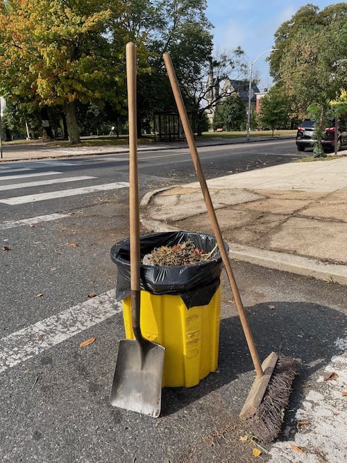 A recycling can full of dirt and leaves with a shovel and brush next to it.