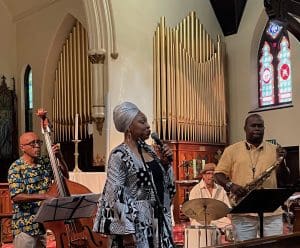 A woman in a headwrap sings into a microphone in front of a band with upright bass, drums, and saxaphone, in a church with a gold organ.