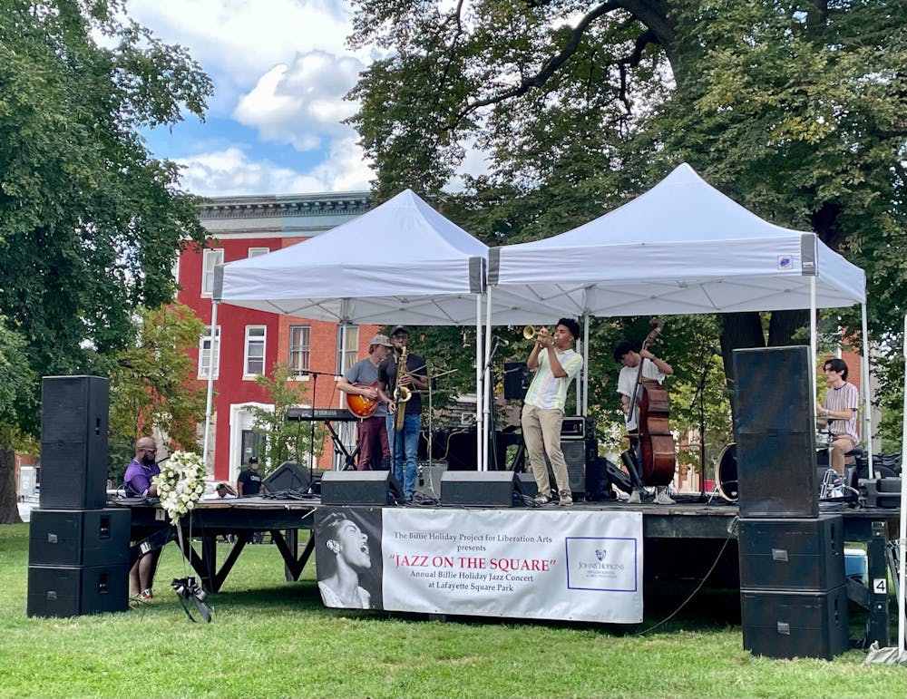 Five person jazz band performing on a black riser stage under two white tents. There are black speakers flanking the stage and there is a white horizontal banner at the front of the stage that reads "The Billie Holiday Project for Liberation Arts presents 'Jazz on the Square' Annual Billie Holiday Jazz Concert at Lafayette Square Park."