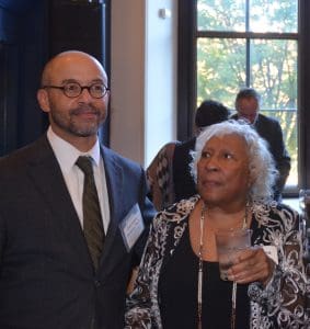 Black man in a black suit with a white button up shirt and striped tie smiling and standing next to a shorter Black woman also in Black dress clothes. She is holding a clear drink and gazing to her right.