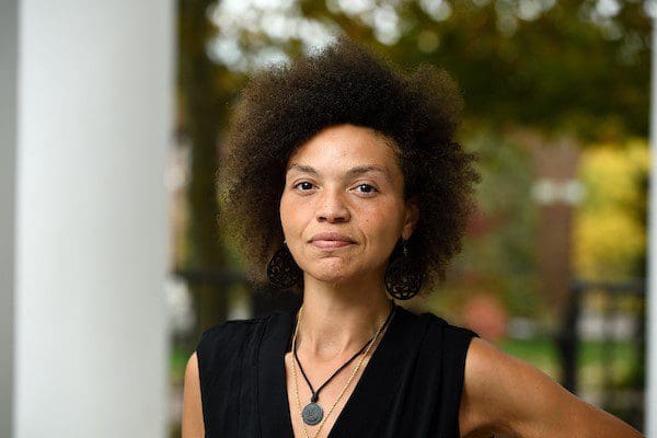 A Black woman gazing intently at the camera while wearing a Black v-neck top with a black rope necklace holding a green stone.