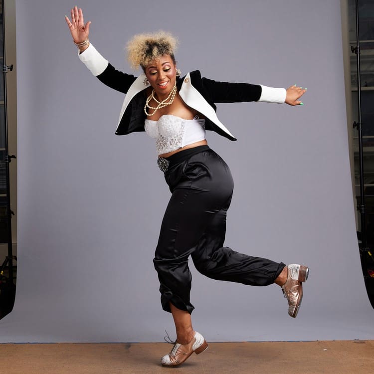 A tap dancer in a black and white outfit posing mid-dance in front of a grey background