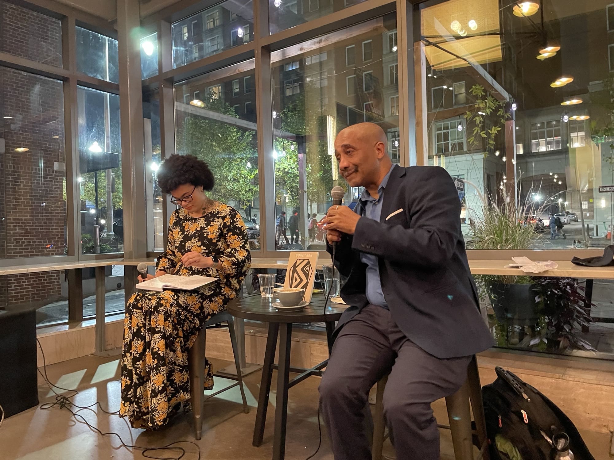 Dr. Lans looking over to David Mills as he holds a microphone and addresses the crowd. A small table sits in between them that holds two cups of water, a cup of coffee, and a copy of his book. 