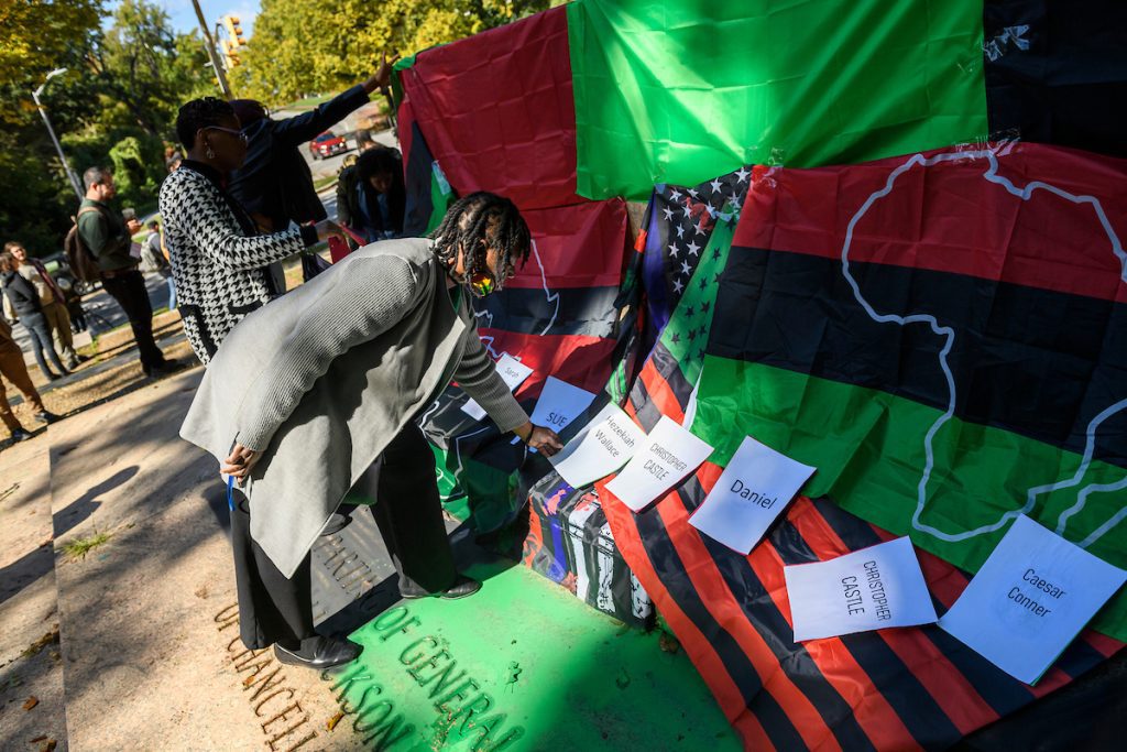 Woman in gray jacket places white sheet of paper with name of enslaved person on red, green, and black flag draped over plinth