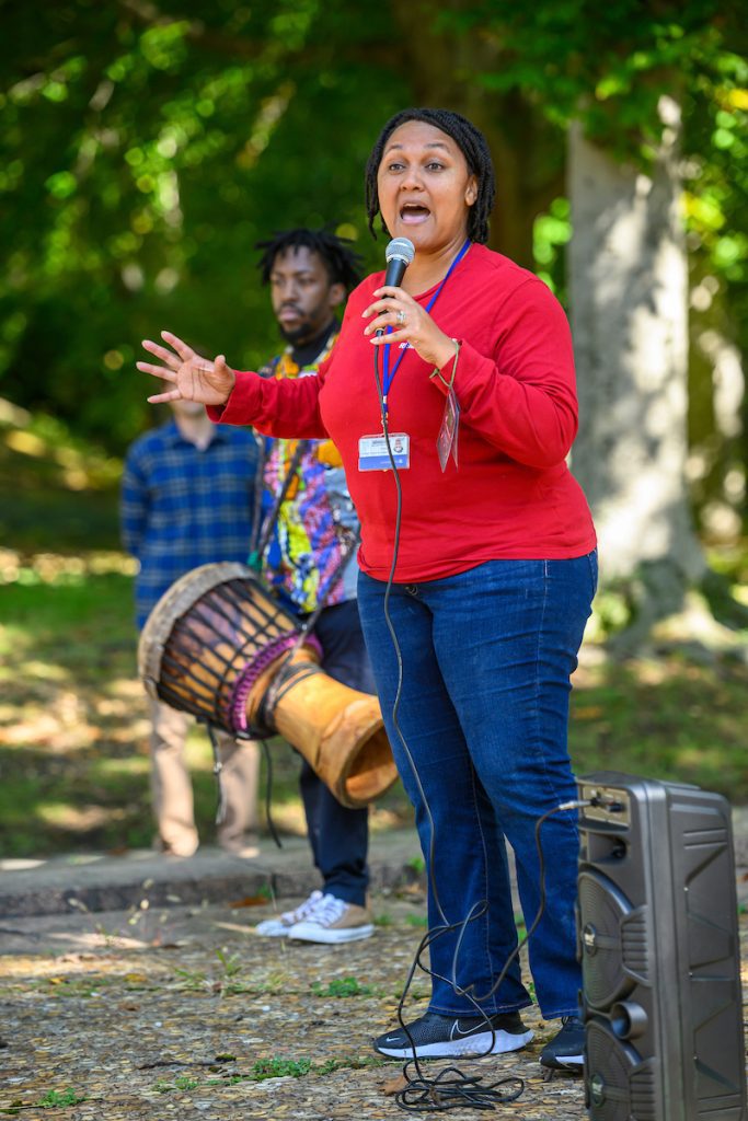Black woman in red shirt and blue jeans speaks into microphone in front of Black man holding drum