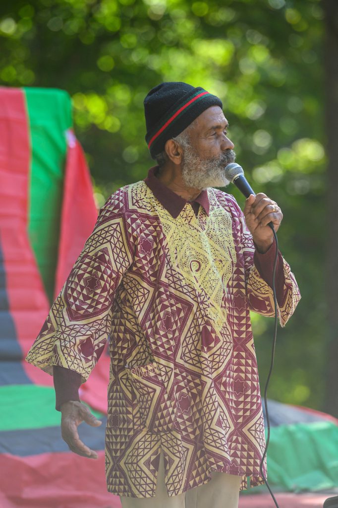 Elder Black man with gray beard in gold and maroon shirt speaks into microphone
