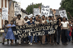 Image of Black protesters with banner and signs marching in the street (from Mangrove film)
