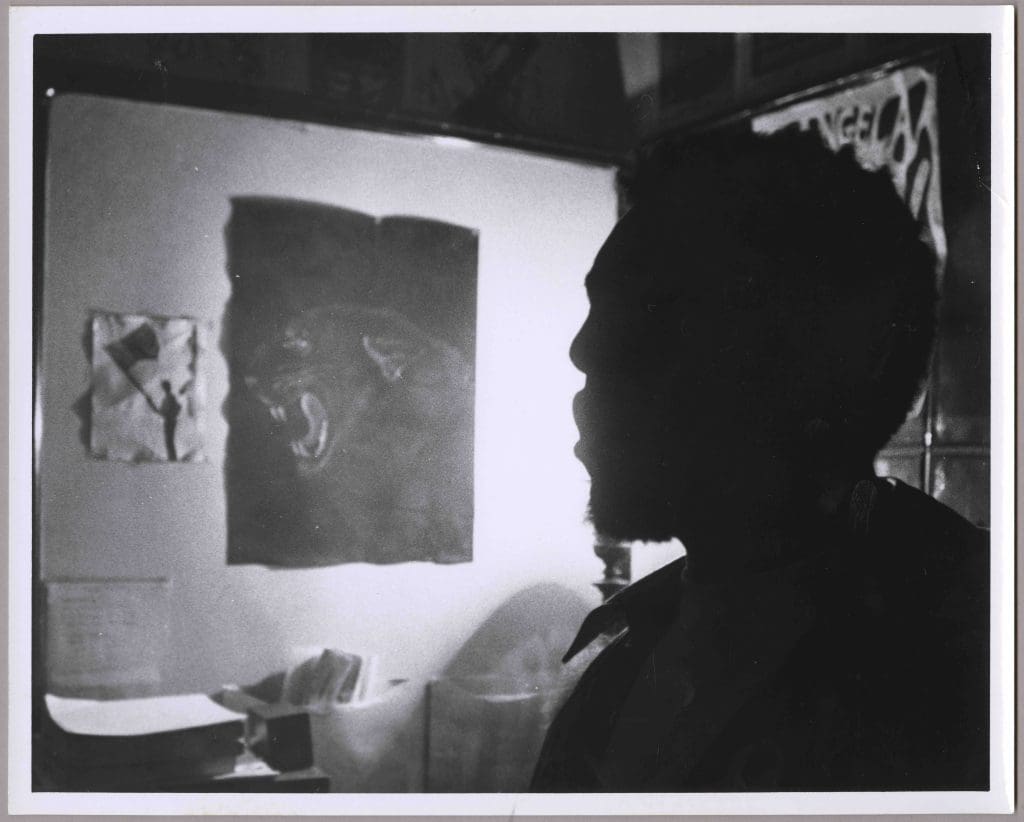 Black Panther Party male member in silhouette in Baltimore headquarters, with Angela Davis, panther, and flag posters behind him.