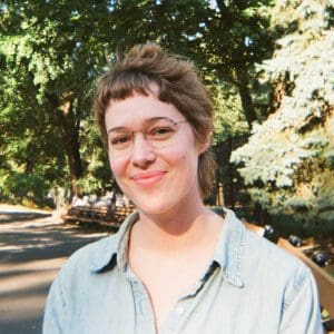 Photo of white woman with glasses and brown hair and denim shirt against leafy trees in background