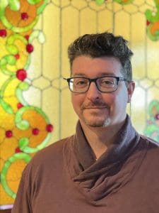 Photo of white man with glasses and goatee and dark hair in brown sweater against yellow stained-glass background