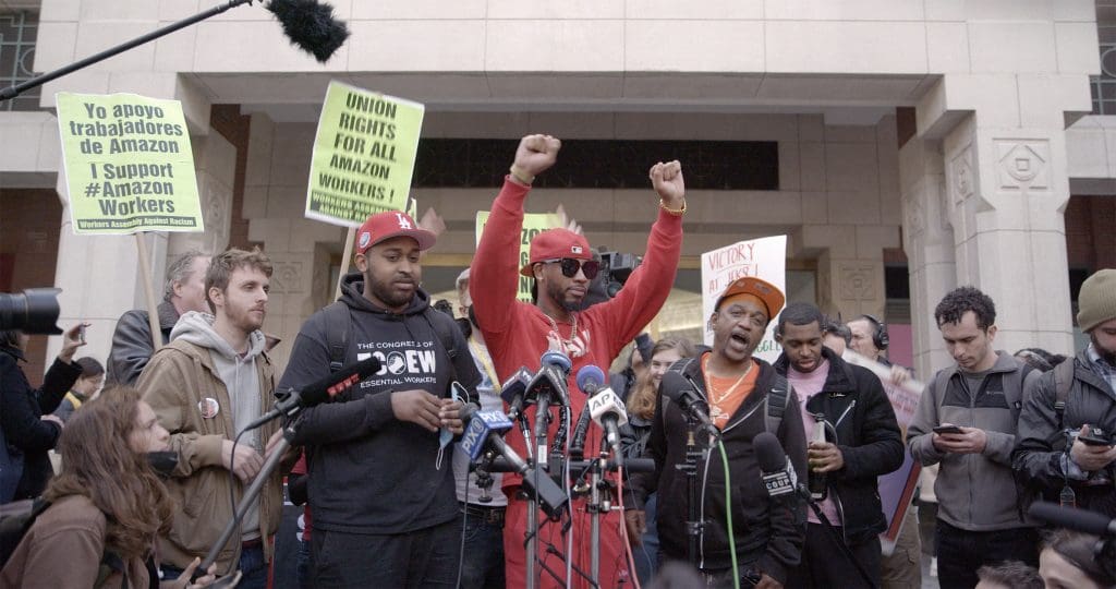 Photo of scrappy union organizers celebrating in front of stately building, including Chris Smalls, Black man with red shirt and hat with fists raised in air